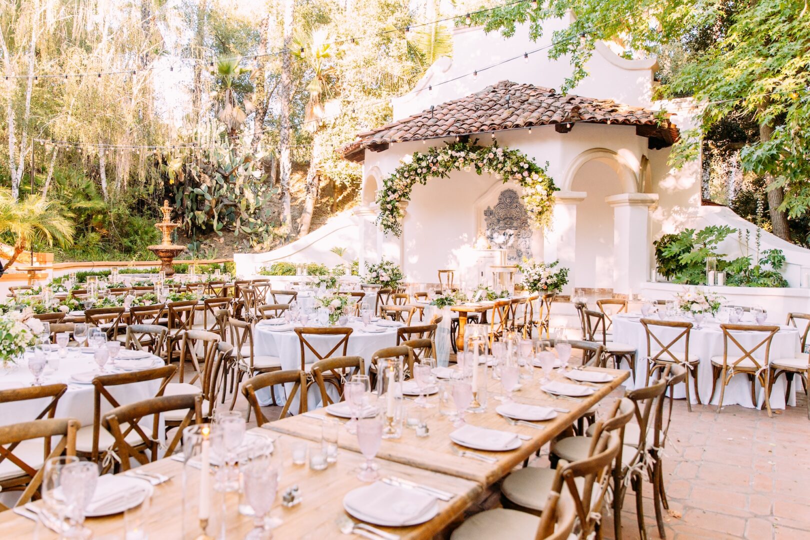 Elegant outdoor wedding reception with long wooden tables, white linens, and floral centerpieces. A small, white, Spanish-style building with a tile roof and floral arch is in the background, surrounded by greenery.