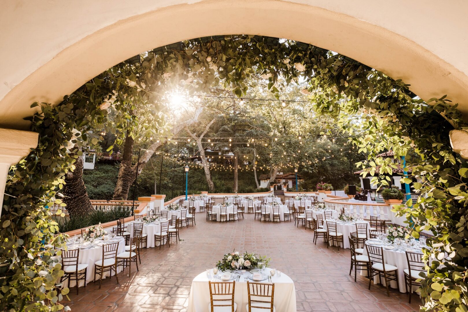 Elegant outdoor wedding reception under an arched entryway with greenery, round tables with white linens, wooden chairs, and string lights hanging above on a sunny day.