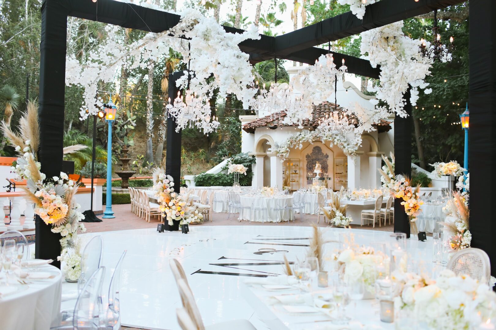 Elegant outdoor wedding reception with white floral decorations, draped fabric, round tables, and a small white building in the background. The setting is bright and festive, surrounded by greenery and soft ambient lighting.