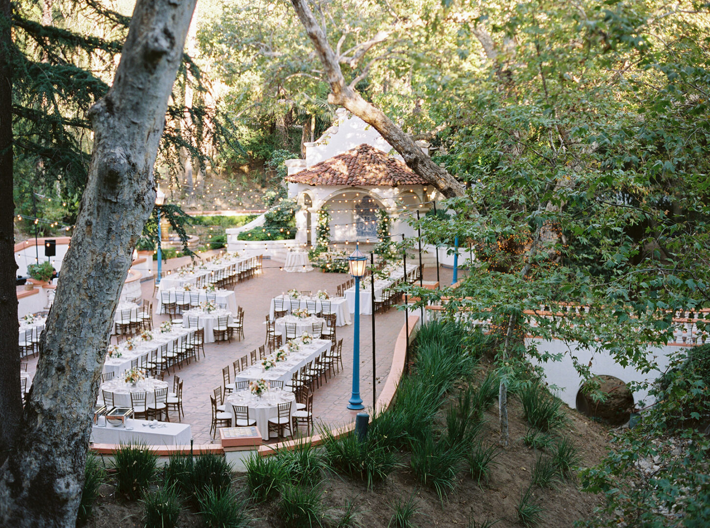 Outdoor wedding venue with long banquet tables, white tablecloths, and chairs arranged on a brick patio near a small pavilion. Lush greenery and tall trees surround the space. String lights are visible above the seating area.