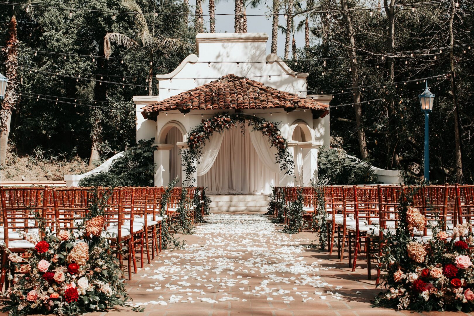 Outdoor wedding venue with rows of wooden chairs, floral arrangements along the aisle, white rose petals on the ground, and a decorated arch in front of a small building with a red-tiled roof.