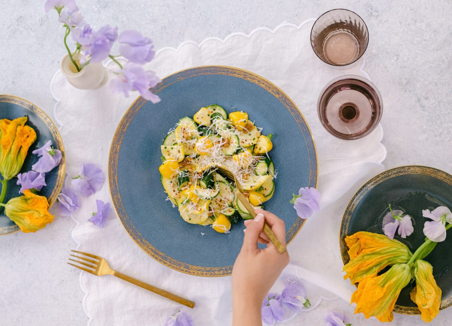 A hand holding a fork over a plate of zucchini and yellow squash pasta, topped with grated cheese. The table is set with flowers, gold utensils, and glasses on a white cloth.