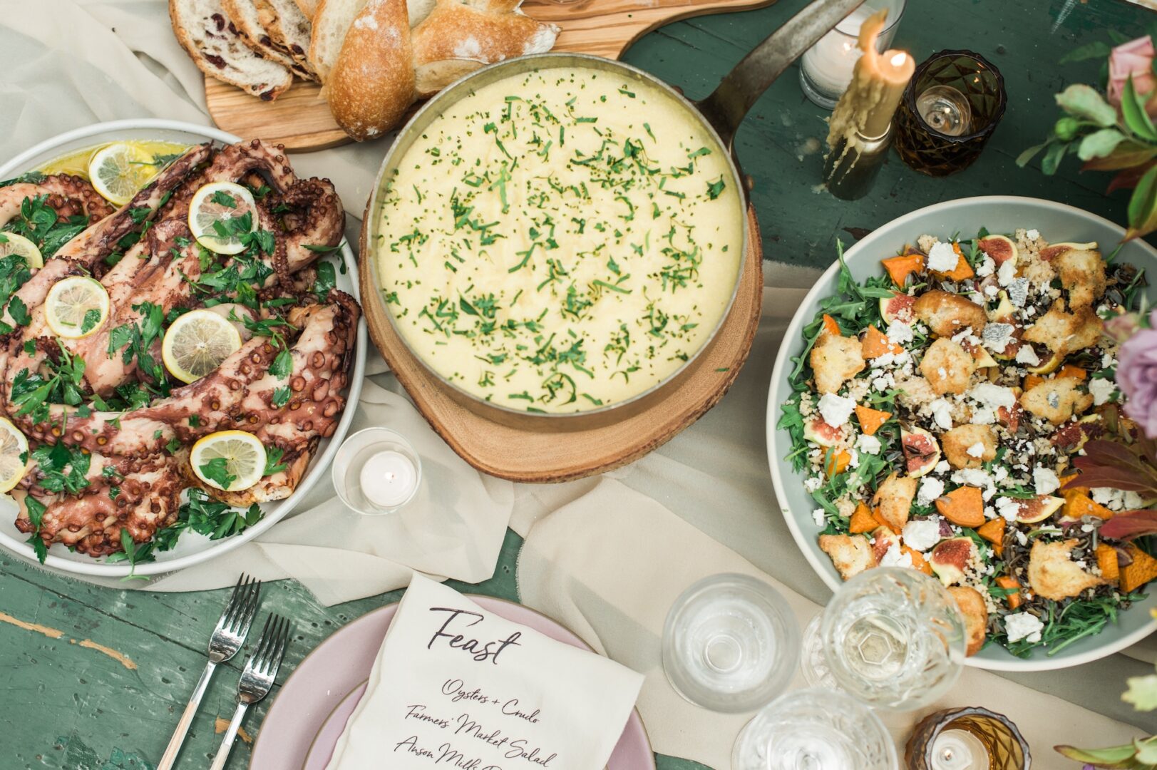 A table set with mashed potatoes, grilled octopus with lemon, a mixed salad with croutons and cheese, bread, candles, glassware, and a handwritten menu on a green and cream tablecloth.