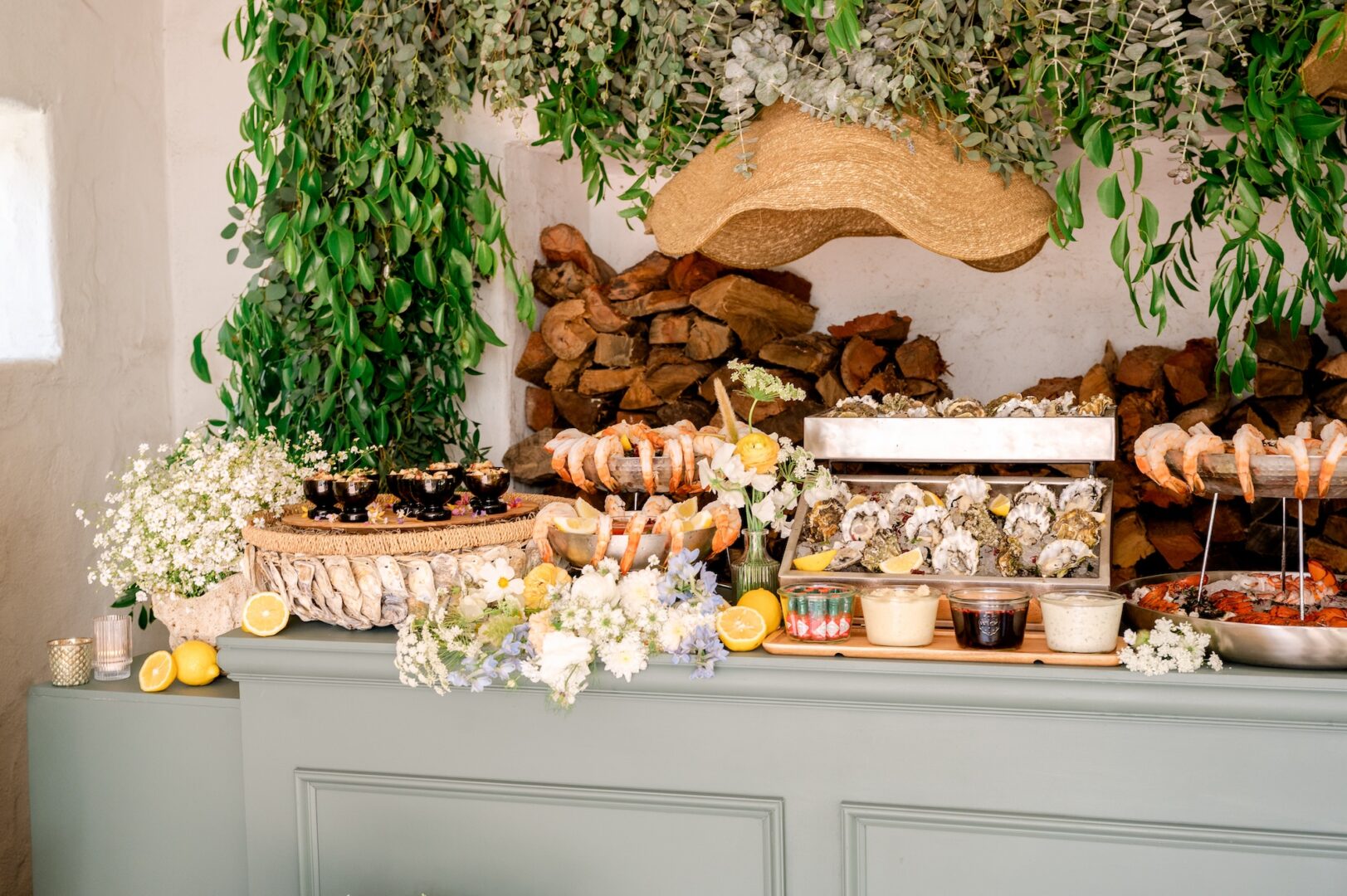 A seafood buffet with oysters, shrimp, sauces, and flowers on a green counter, surrounded by leafy greenery and stacked wood in the background.