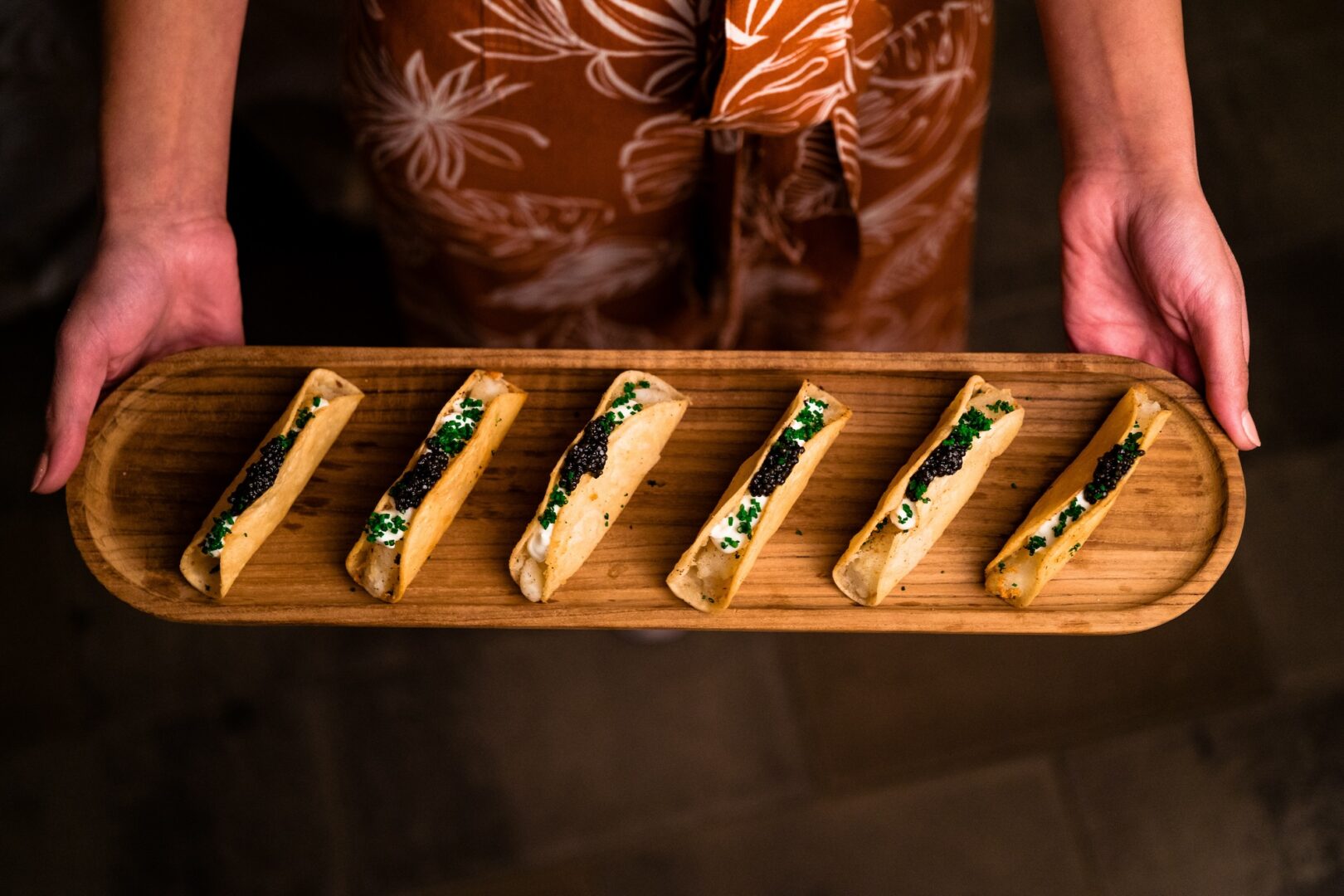 A person holds a wooden tray with six neatly arranged, bite-sized appetizers, topped with green herbs and a dark garnish. The person wears a brown floral-patterned outfit.