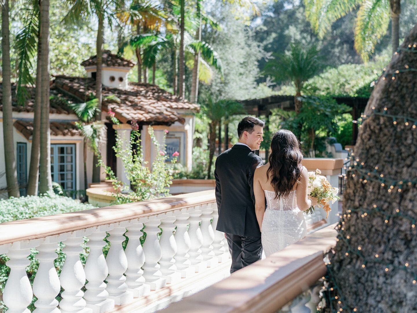 A bride and groom walk together on a sunlit balcony surrounded by lush greenery and palm trees, with a Mediterranean-style building in the background.