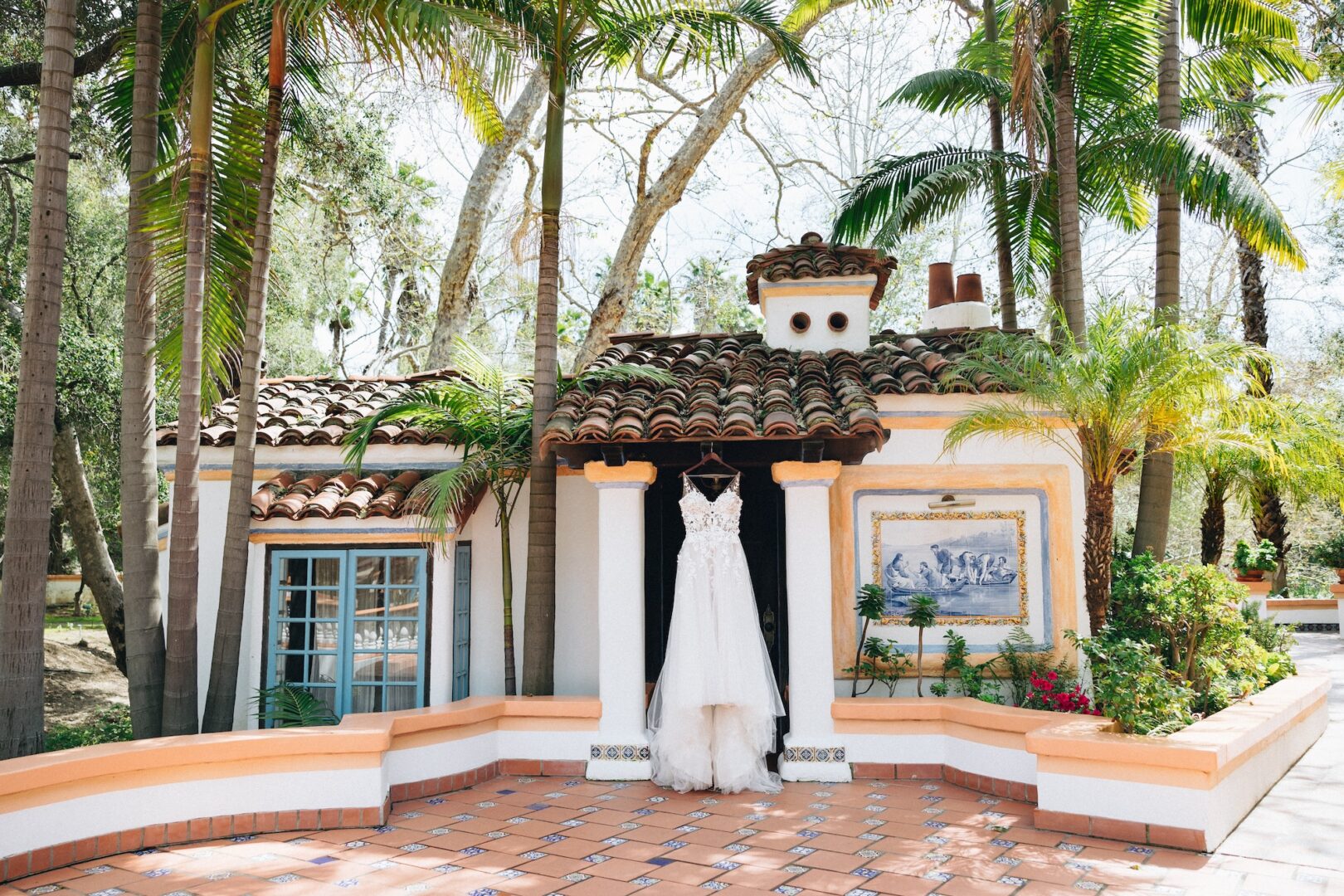 A white wedding dress hangs outside a small, Spanish-style villa with terracotta roof tiles, palm trees, and colorful tile details in a sunny outdoor setting.