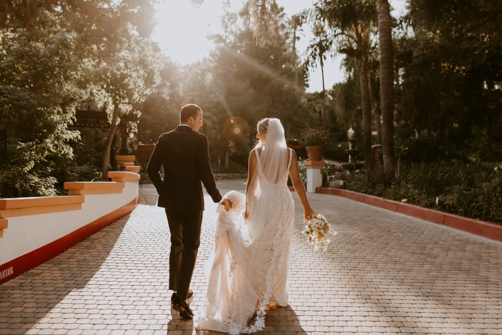 A bride and groom walk hand in hand down a sunlit, tree-lined path. The bride holds a bouquet and her dress train, while warm sunlight filters through the trees, creating a romantic atmosphere.
