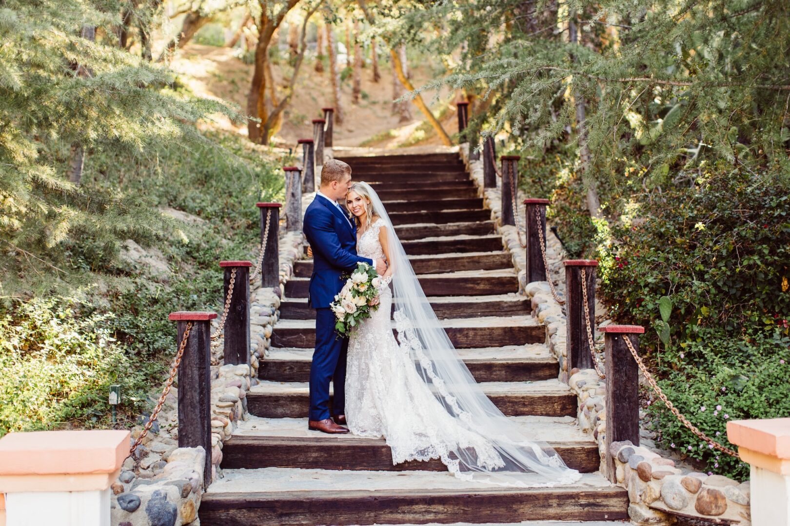 A bride in a white gown and veil stands with a groom in a blue suit on outdoor stone steps, surrounded by lush greenery and trees, smiling and embracing.
