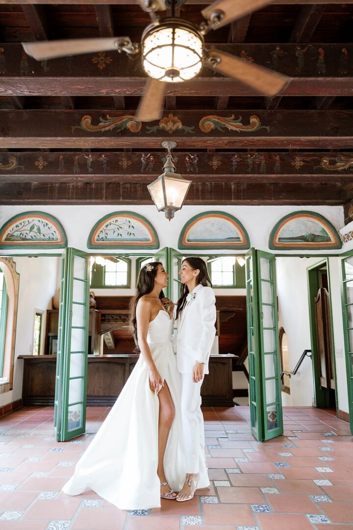 Two women in white wedding outfits stand facing each other and smiling in a sunlit room with green doors, wooden ceiling beams, and decorative tiles.