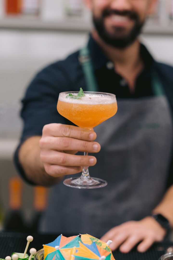 A bartender in a gray apron holds out an orange cocktail in a coupe glass, garnished with mint. The person is smiling, and a colorful drink umbrella is visible in the foreground.
