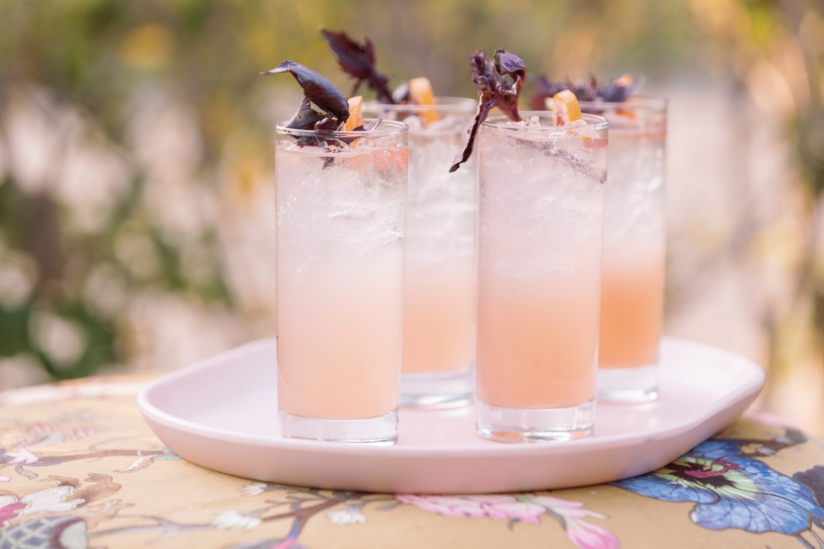 Four tall glasses of a light pink drink with ice are garnished with dark purple leaves and a small orange curl, arranged on a white tray outdoors.