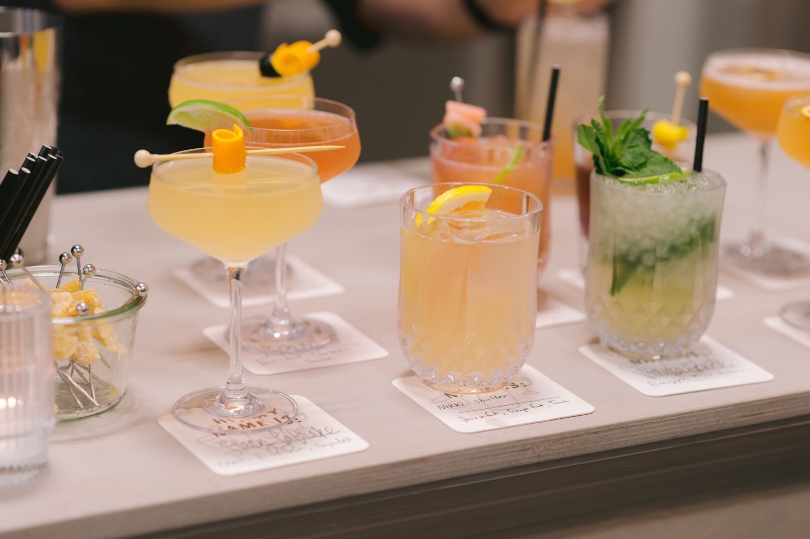 A row of colorful cocktails in different glasses, each garnished with fruit, herbs, or skewers, sits on a bar counter with napkins underneath each drink.