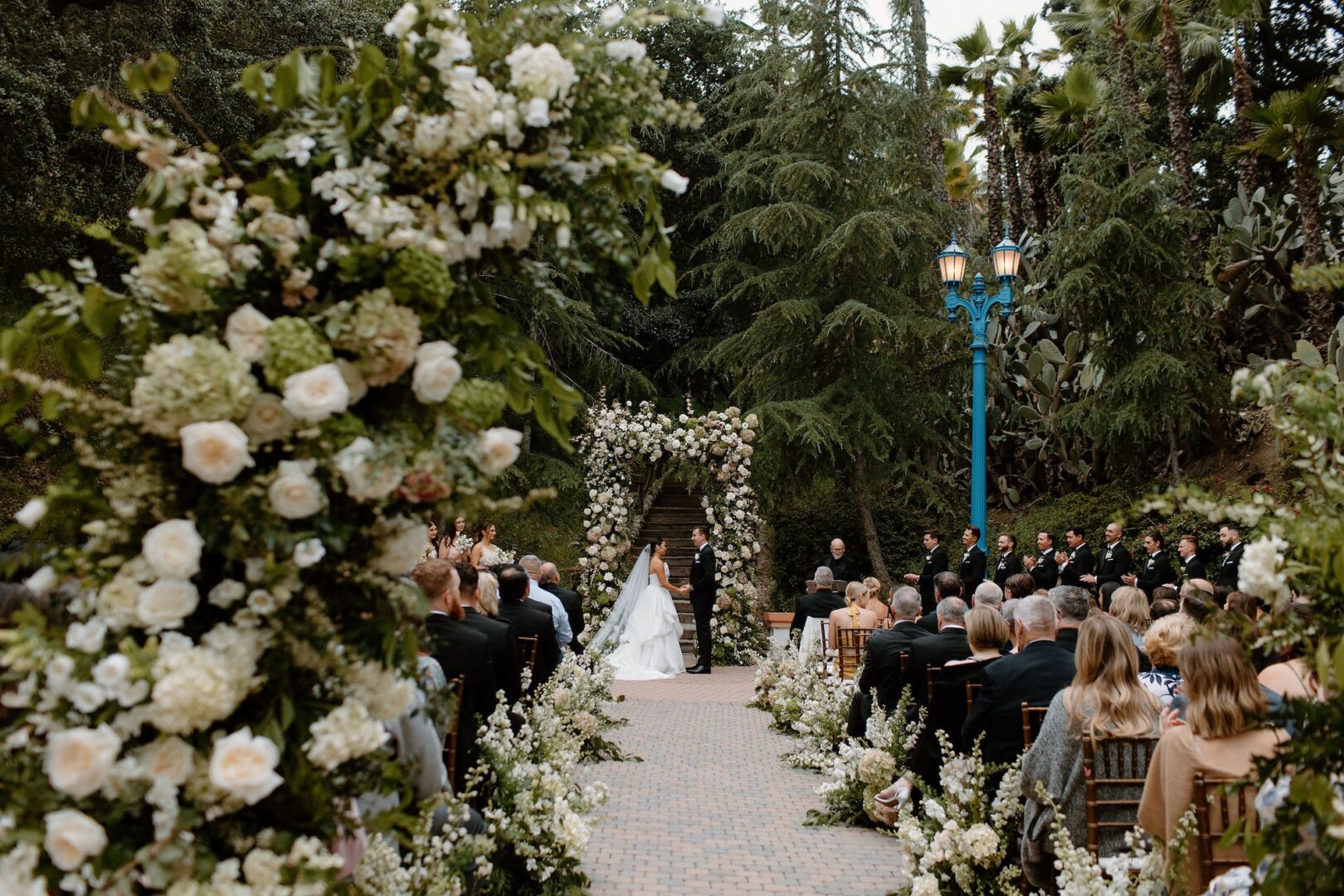 An outdoor wedding ceremony with a bride and groom standing under a floral arch, surrounded by seated guests. Lush greenery and white flowers decorate the aisle. Trees line the background, and the scene appears elegant and serene.