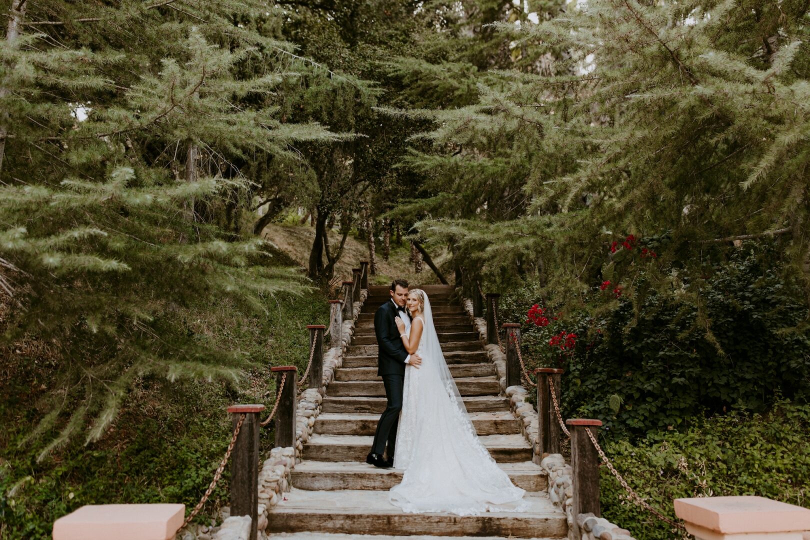 A bride and groom embrace on stone steps surrounded by lush green trees and foliage, creating a romantic outdoor wedding scene.