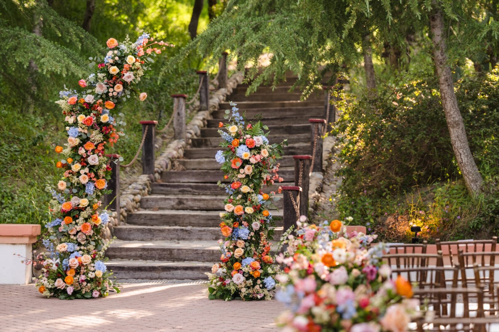 Outdoor wedding setup with wooden steps, lush greenery, and tall flower arrangements in pink, orange, blue, and cream. Rows of chairs and more floral displays create a festive, elegant atmosphere.