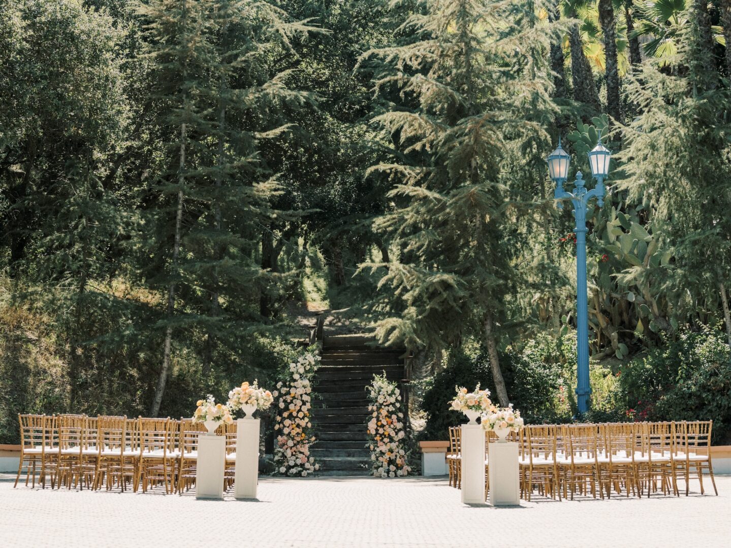 Outdoor wedding ceremony setup with rows of wooden chairs facing floral arrangements and a staircase, surrounded by tall green trees and a blue lamp post in the background.