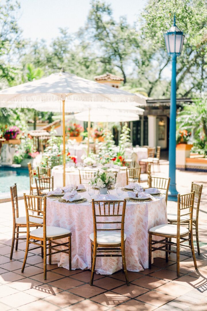 A round table set for an outdoor event with eight wooden chairs, a white floral tablecloth, elegant table settings, centerpiece flowers, and a large white parasol in a sunny garden patio.