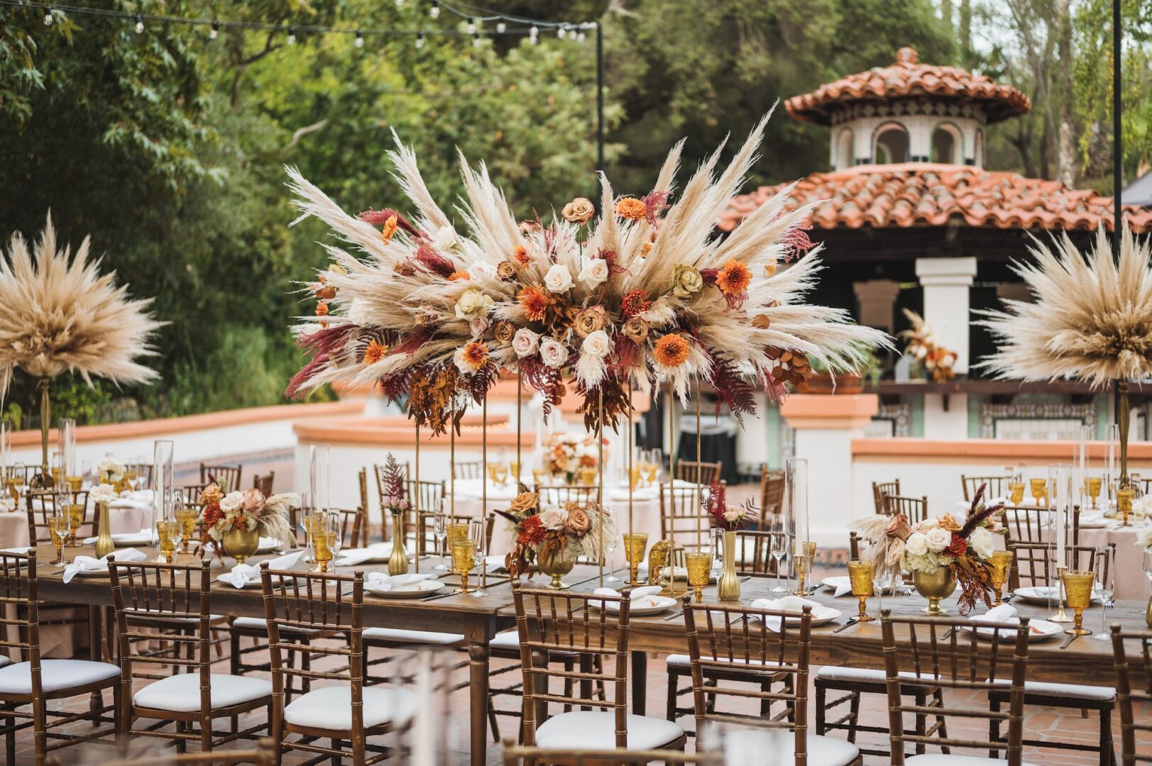Outdoor event setup with wooden tables, brown chairs, and tall floral centerpieces of pampas grass and autumn flowers. Background shows greenery and a building with a red tile roof. Festive, elegant atmosphere.