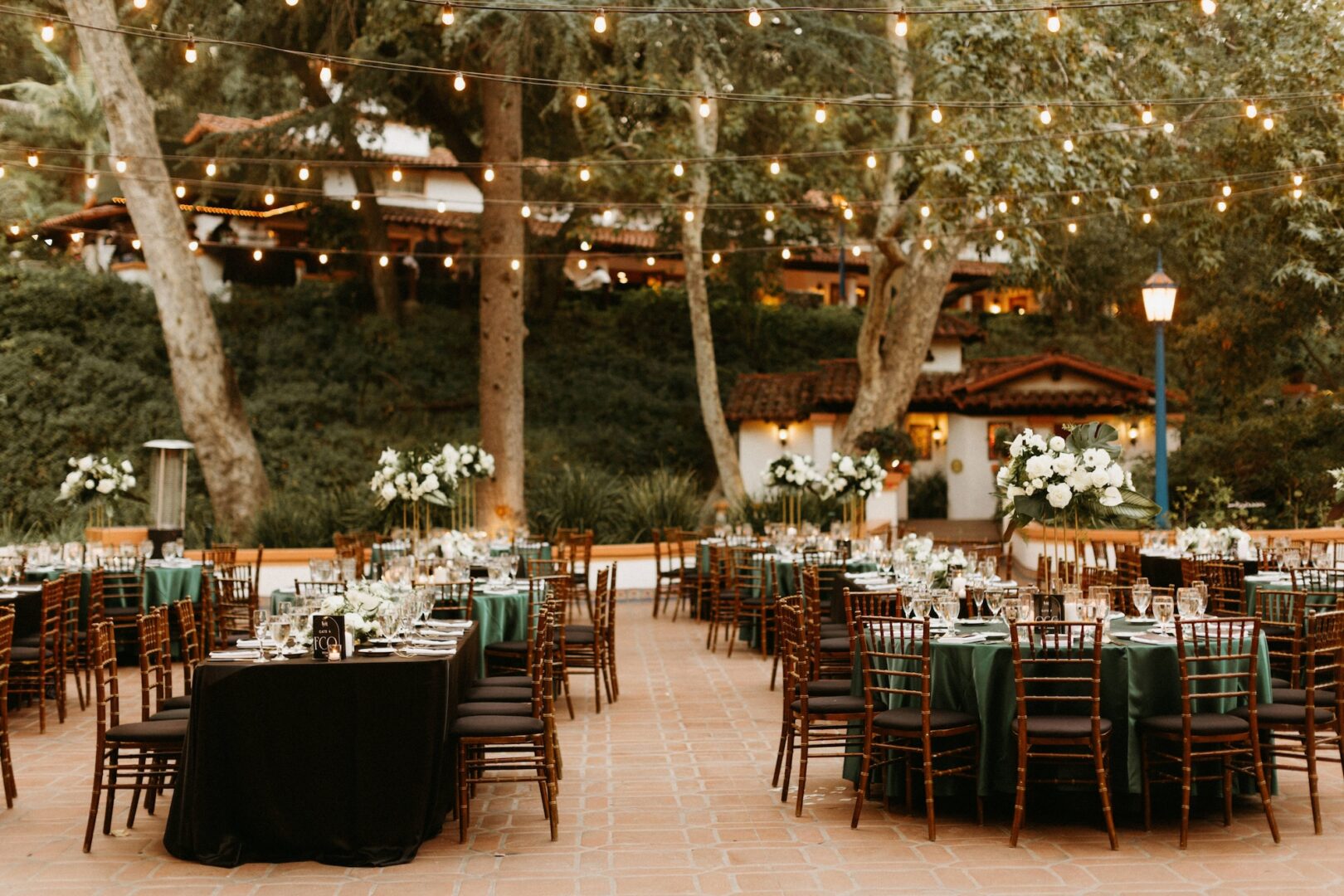 Outdoor wedding reception setup with round tables, green tablecloths, elegant floral centerpieces, wooden chairs, and string lights overhead. Trees and buildings create a cozy, festive atmosphere in the background.