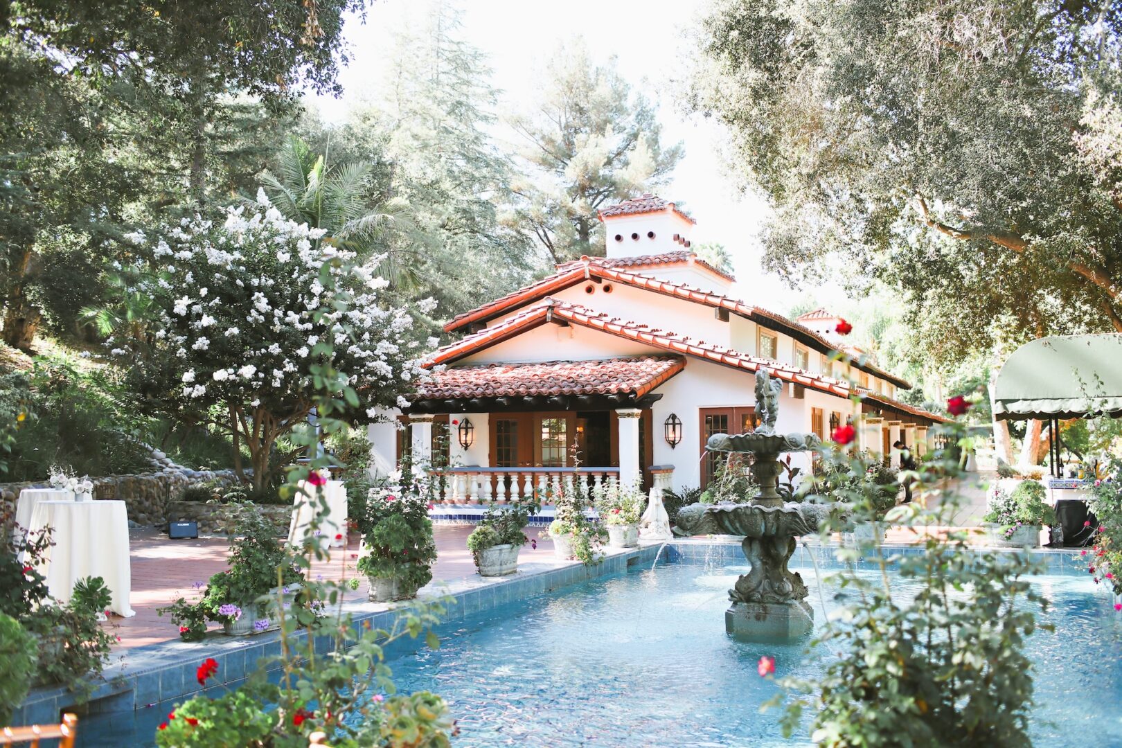 A bright villa with a red-tiled roof sits behind a garden fountain, surrounded by lush trees, blooming flowers, and a clear blue pool on a sunny day.