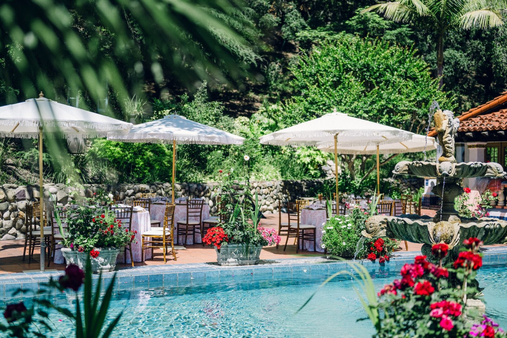 Tables and chairs with white umbrellas are set up beside a swimming pool surrounded by greenery, potted flowers, and a stone fountain, creating an inviting outdoor event setting.