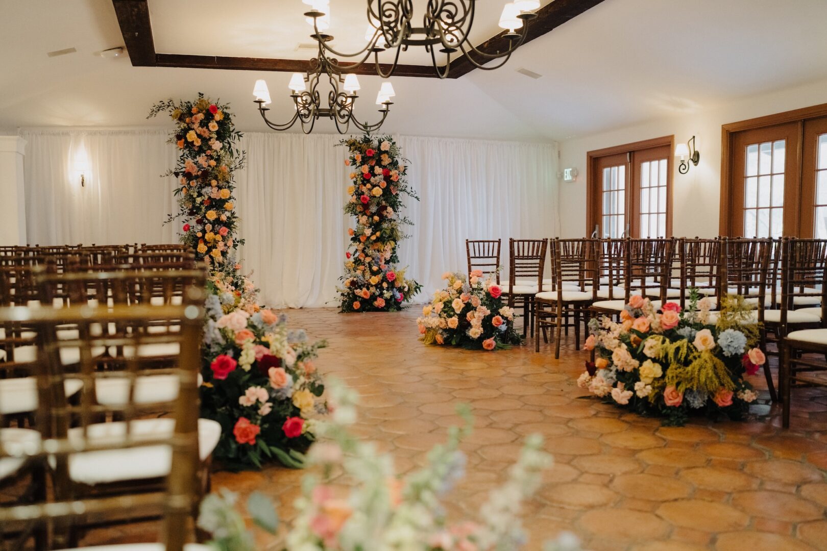 Indoor wedding ceremony setup with rows of wooden chairs, floral arrangements lining the aisle, and two tall flower pillars in front of a white draped backdrop, under elegant chandeliers.