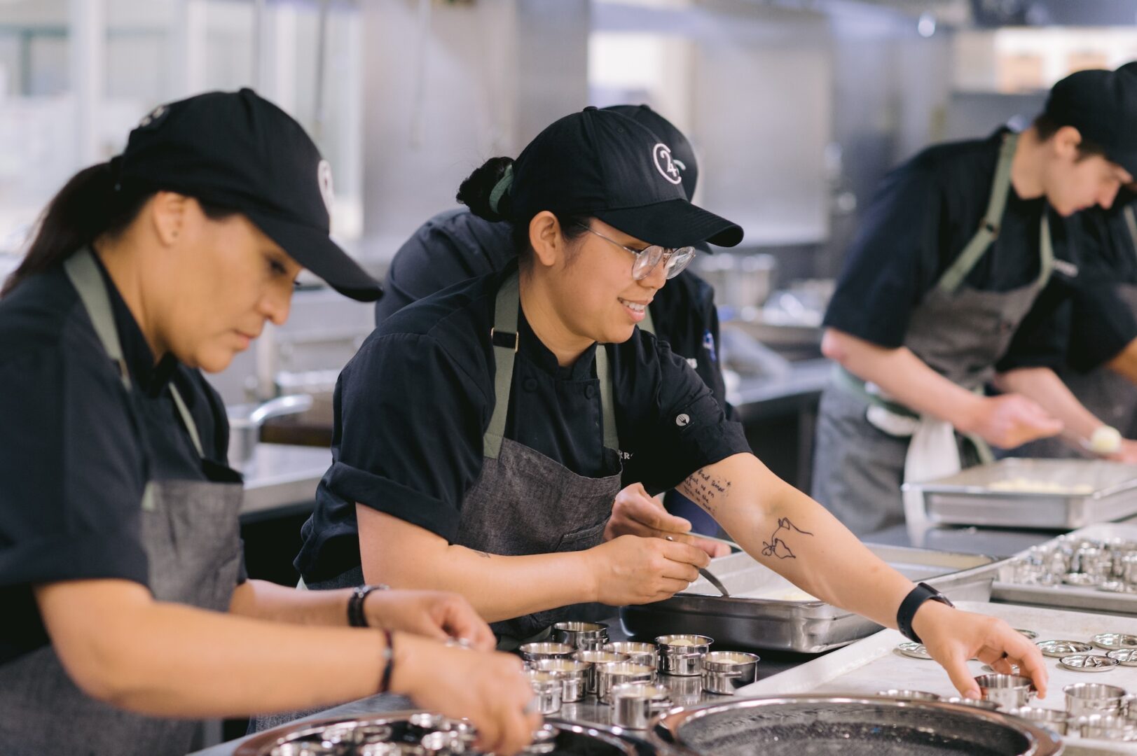 Three chefs in black uniforms and gray aprons prepare food in a commercial kitchen, focusing intently on their tasks. One chef in the center is smiling while arranging metal containers.