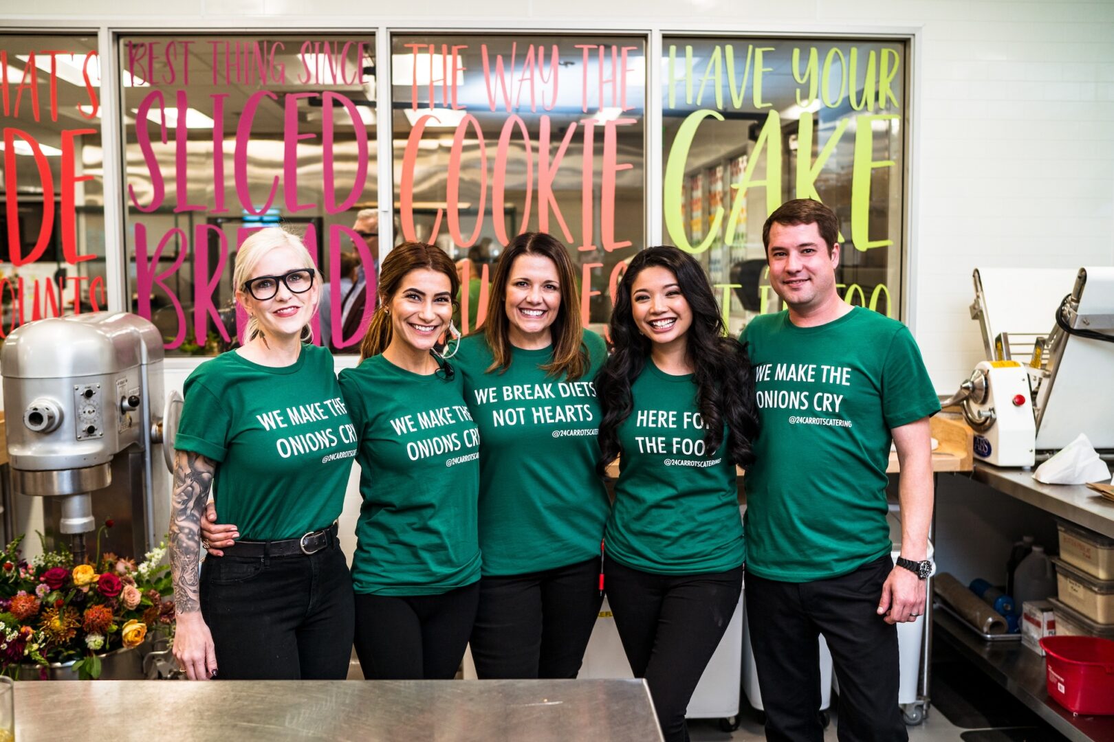 Five people wearing green slogan t-shirts stand side by side in a kitchen, smiling. Colorful text decorates the glass behind them. Kitchen equipment and flowers are visible in the background.