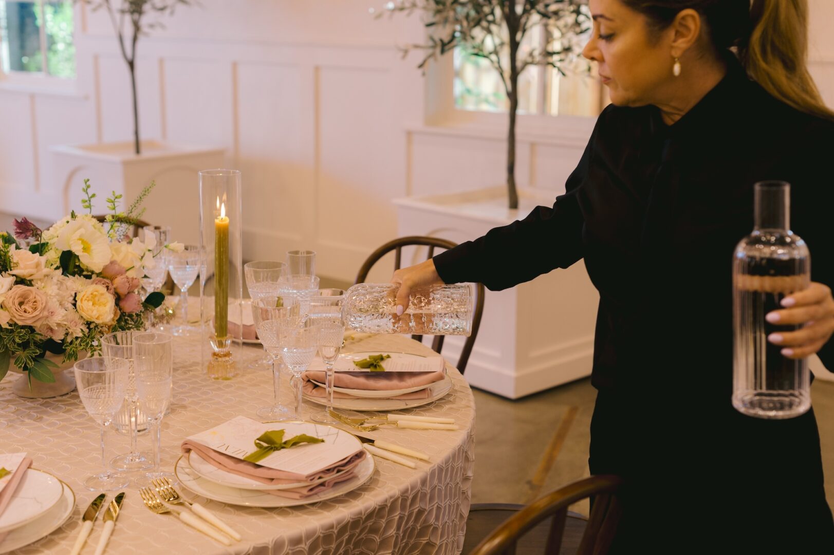 A person in black pours water into a glass at an elegantly set round table with floral centerpiece, candles, and pink napkins in a softly lit room.
