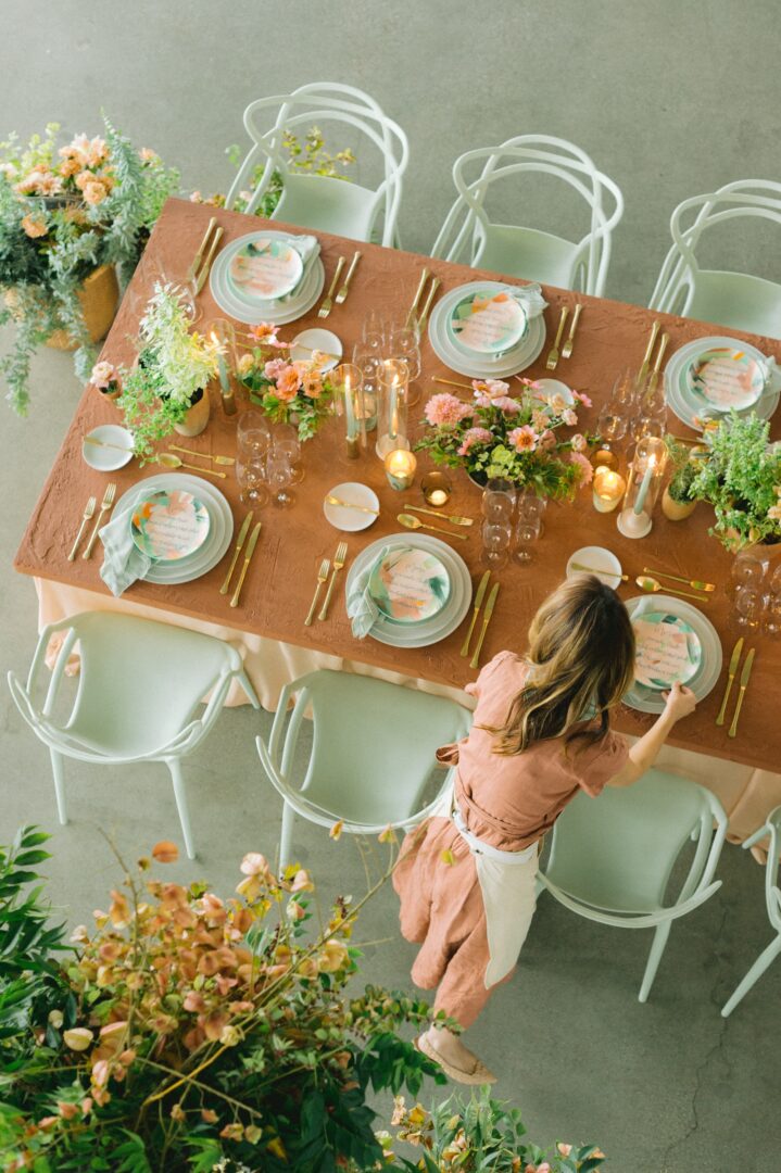 A woman arranges a beautifully set table with pastel plates, gold cutlery, candles, and floral centerpieces. The scene is viewed from above, creating a warm and inviting atmosphere.