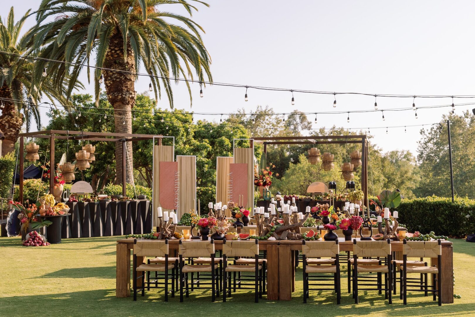 Outdoor event setup with wooden tables and chairs, colorful floral centerpieces, decorative lighting, fruit displays, and palm trees in the background under a clear sky.