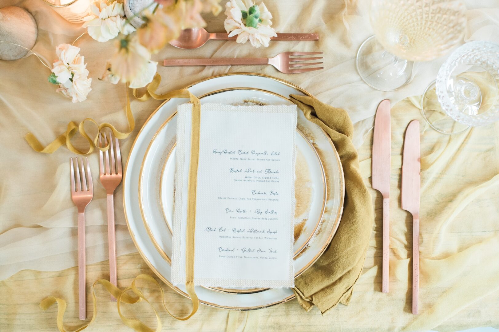 Elegant table setting with gold-rimmed plates, pink cutlery, a fabric menu, flowers, and soft yellow napkins, arranged on a cream and gold cloth. Glassware and candles add a touch of sophistication.