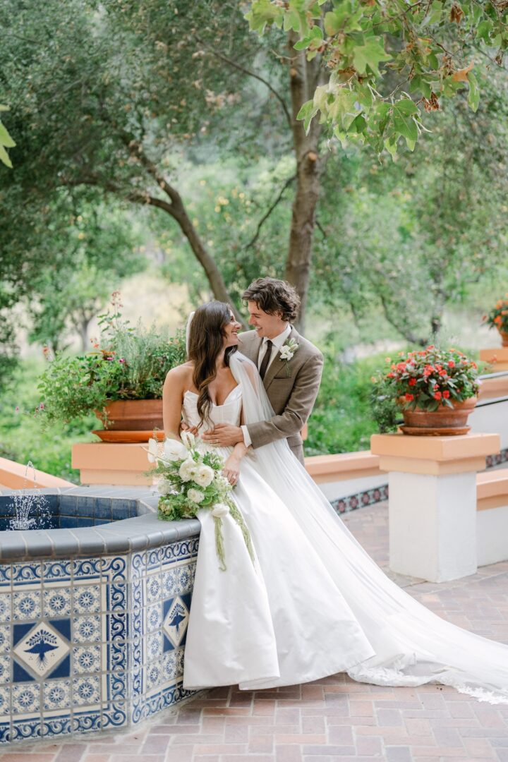 A bride and groom stand by a blue-tiled fountain, surrounded by greenery and flowers. The bride wears a white dress and holds a bouquet, while the groom stands behind her, embracing her and smiling.