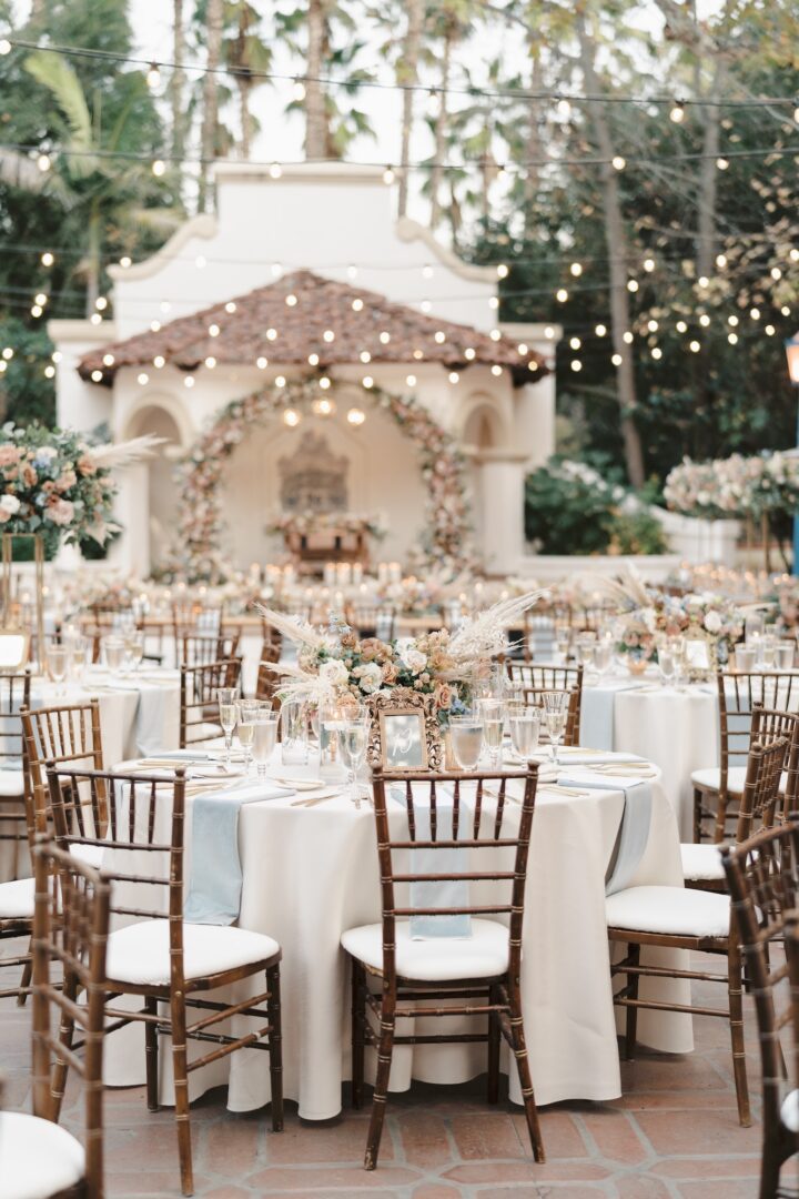 Elegant outdoor wedding reception with round tables, wooden chairs, floral centerpieces, string lights overhead, and a floral arch in front of a white stucco structure surrounded by trees.