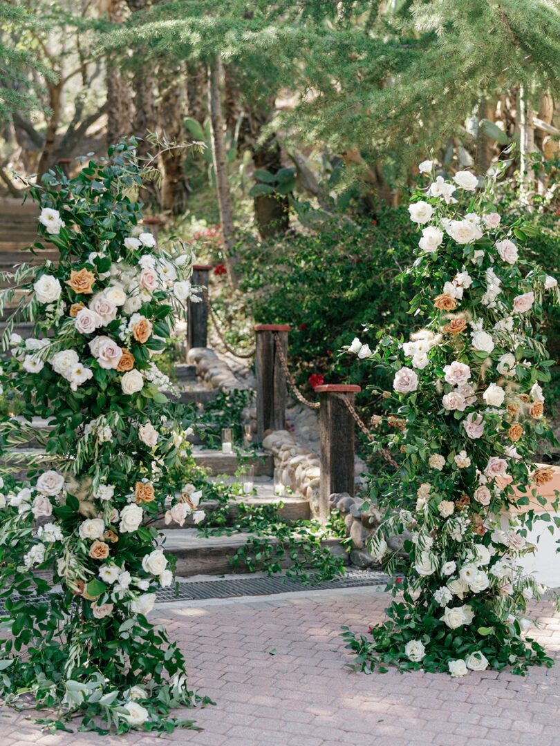 Two large floral arrangements with white and pale pink roses and greenery frame a stone path with steps, leading into a lush garden setting.