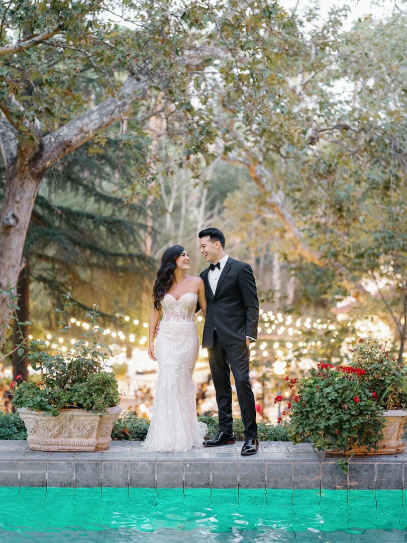 A couple dressed in formal attire stands on the edge of a pool, gazing at each other, with trees and string lights in the background. The scene is romantic and outdoor.