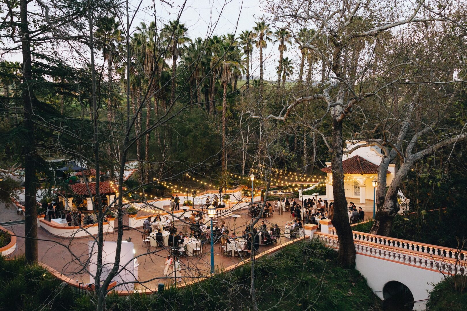 An outdoor event with string lights, people dining at tables, and a white building surrounded by trees and palm trees at dusk.