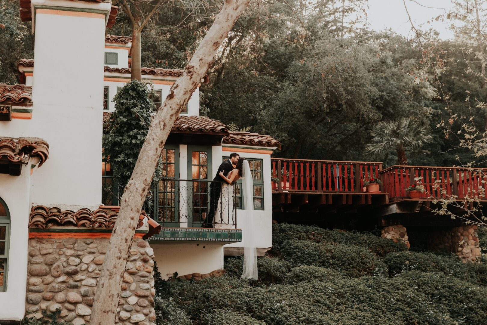 A couple in wedding attire embraces on a balcony of a rustic house surrounded by greenery and trees.