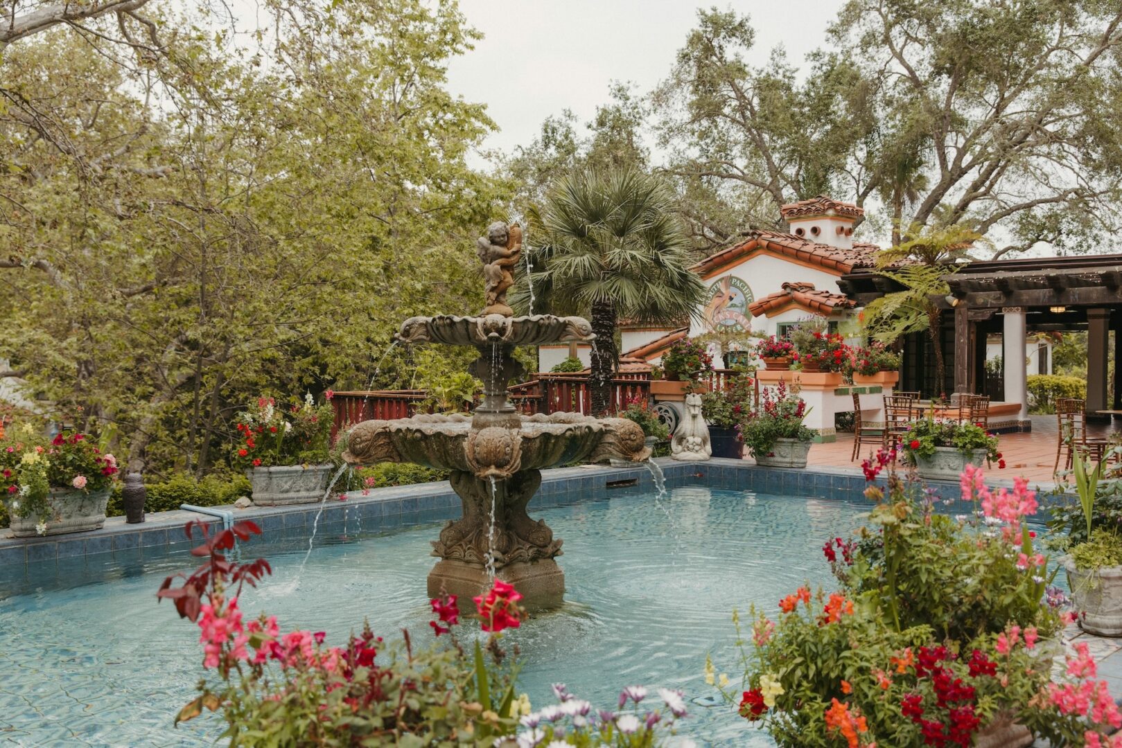 A stone fountain with sculptures stands in the center of a blue pool, surrounded by vibrant flowers, palm trees, and a Spanish-style building with a tiled roof in the background.