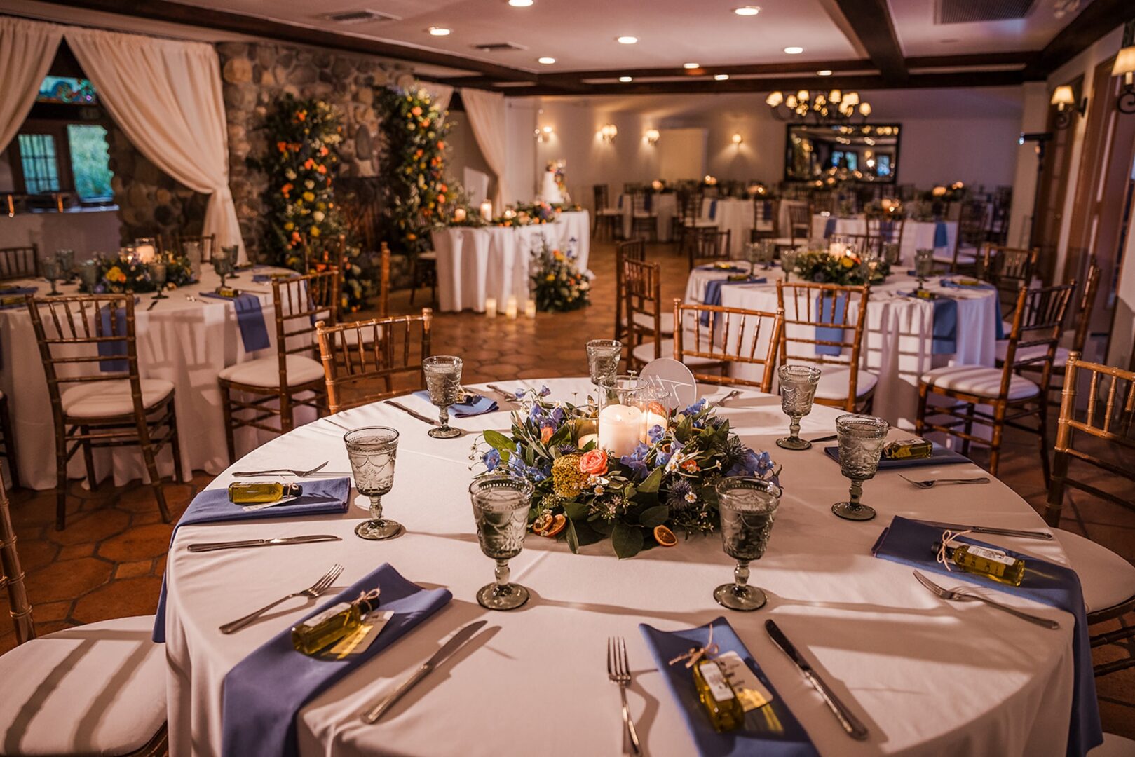 Elegant banquet hall with round tables set for an event, featuring white tablecloths, blue napkins, floral centerpieces with candles, glassware, and small gifts at each place setting. Warm lighting creates a cozy atmosphere.