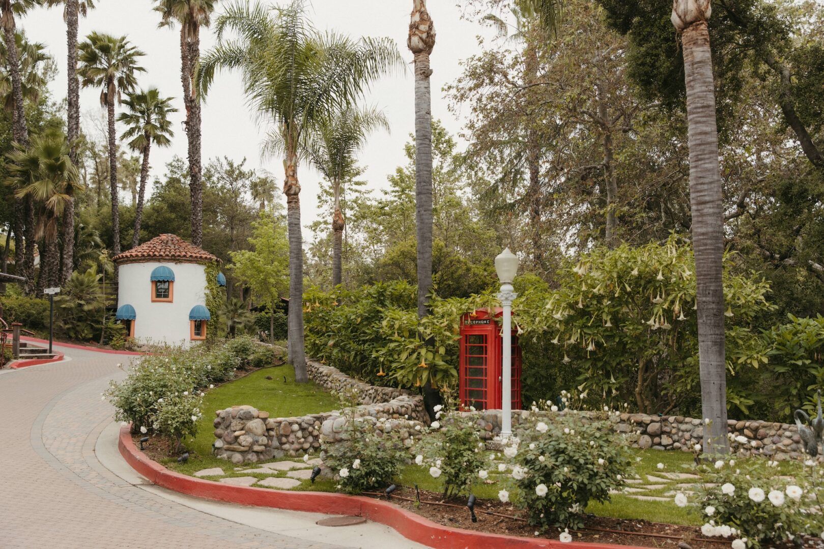A red British-style phone booth stands near a small round white building with blue windows, surrounded by palm trees, greenery, and a curving brick path.