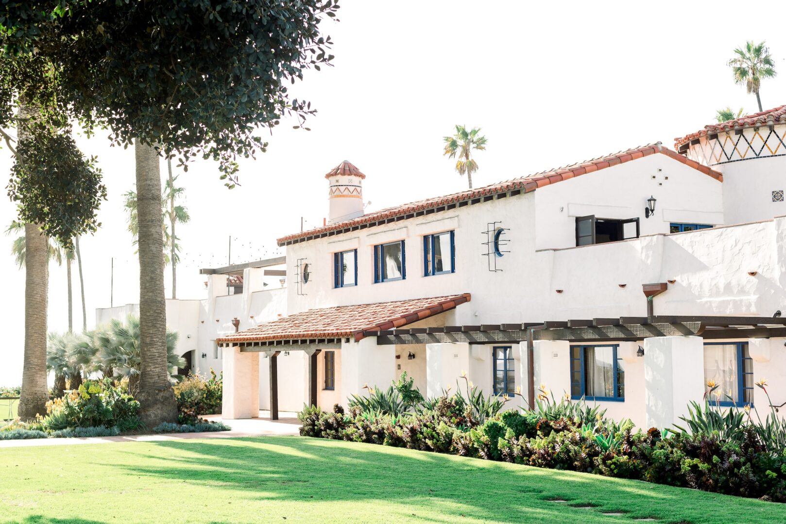 A white stucco building with terracotta roof tiles, surrounded by lush green grass, palm trees, and landscaped plants, under a bright, sunny sky.