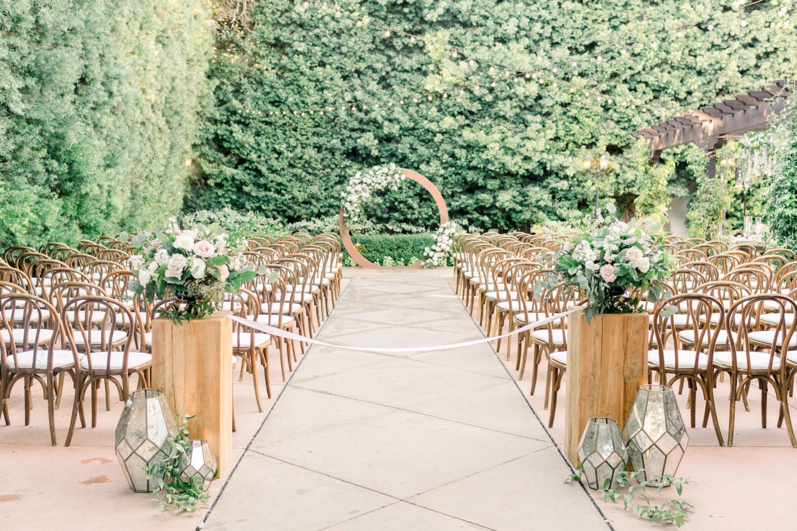 Outdoor wedding ceremony setup with rows of wooden chairs, a flower-decorated circular arch, lush greenery, floral arrangements, and glass lanterns lining the aisle.