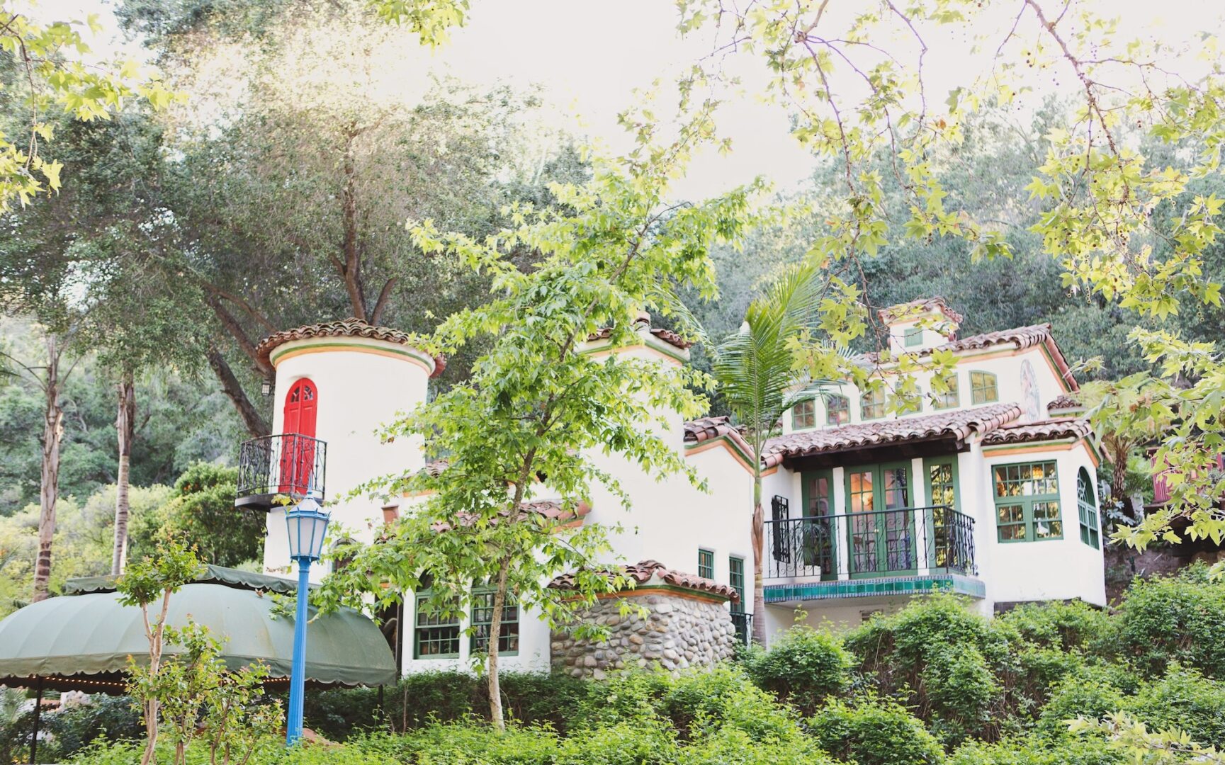 A charming house with white walls, a red door, and stone accents nestled among trees and greenery, featuring balconies and a turret, in bright, natural daylight.