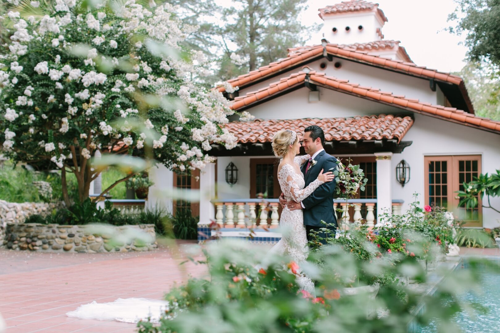 A bride and groom embrace in front of a Spanish-style house, surrounded by greenery and flowers, on their wedding day.
