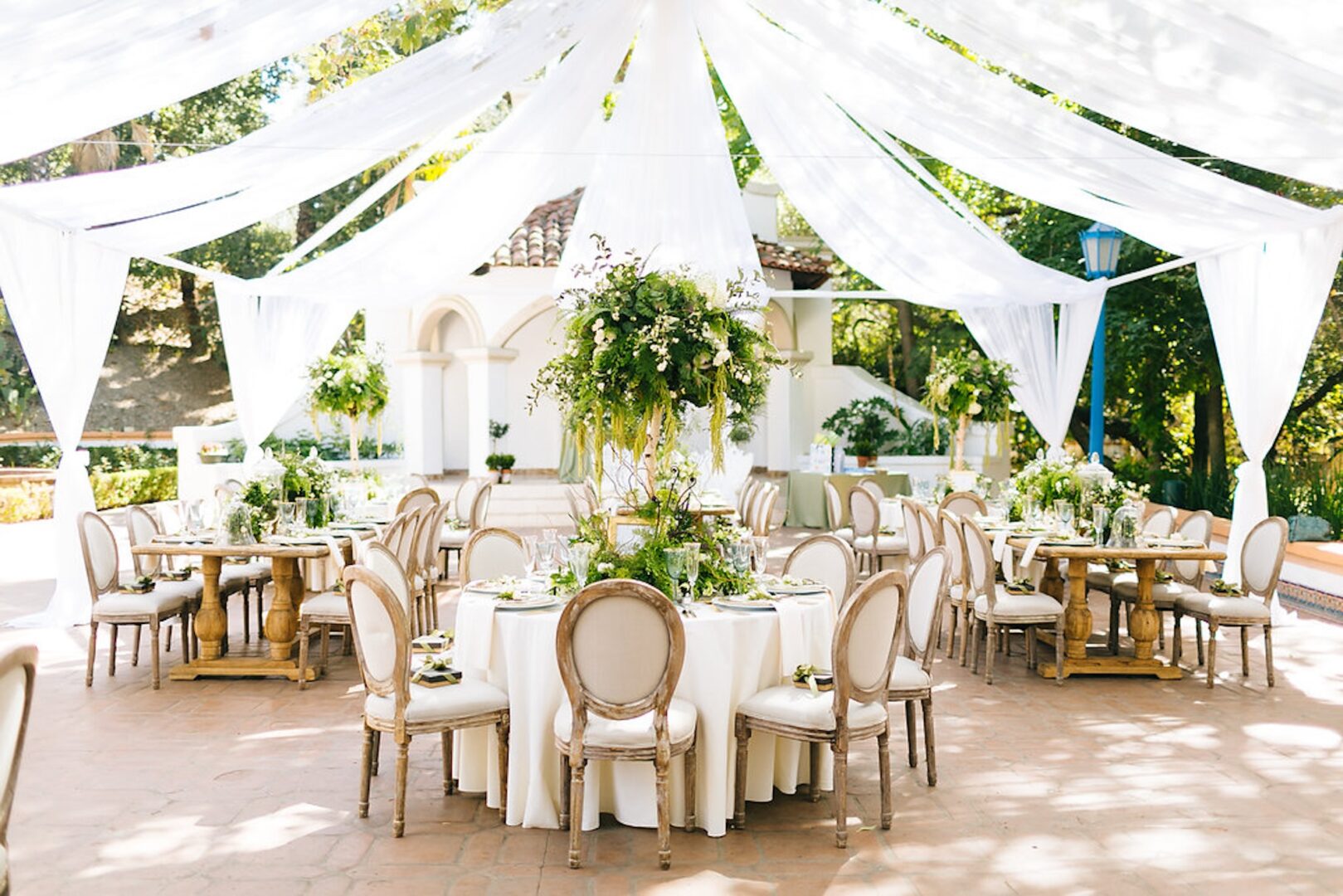 Elegant outdoor event with round tables, wooden chairs, white tablecloths, and green floral centerpieces under flowing white drapes, surrounded by greenery and sunlight.