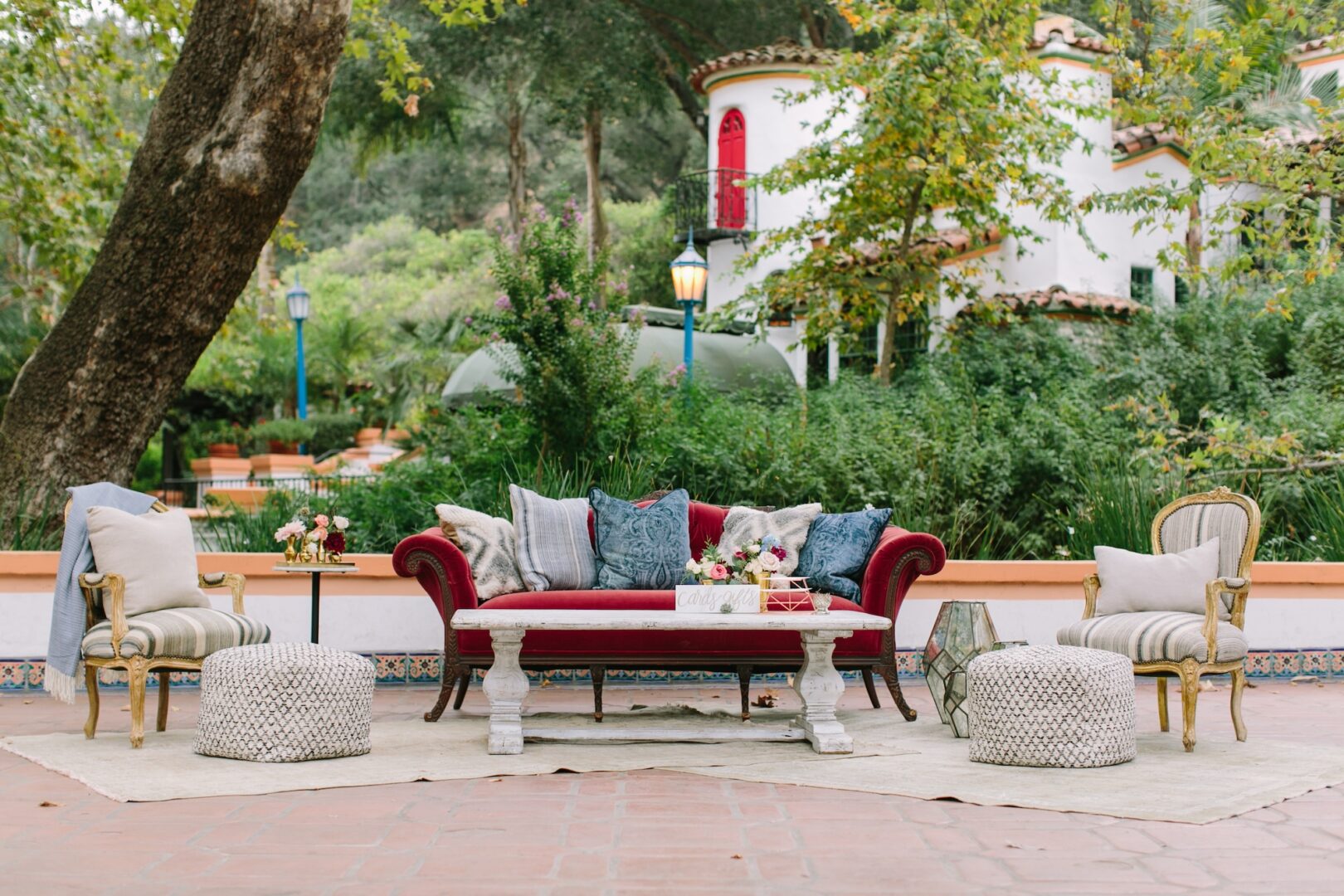 Outdoor seating area with a red velvet sofa, cozy pillows, round ottomans, armchairs, a white coffee table, and side tables with flowers, set on a rug. Lush greenery and a white house with red trim are in the background.