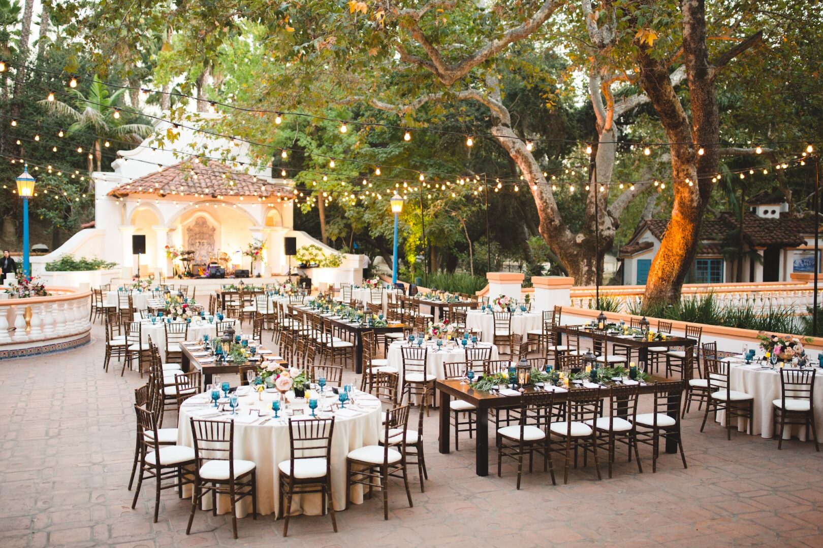 Outdoor wedding reception setup with round and rectangular tables, brown chairs, floral centerpieces, and string lights under large trees, with a decorated stage in the background.
