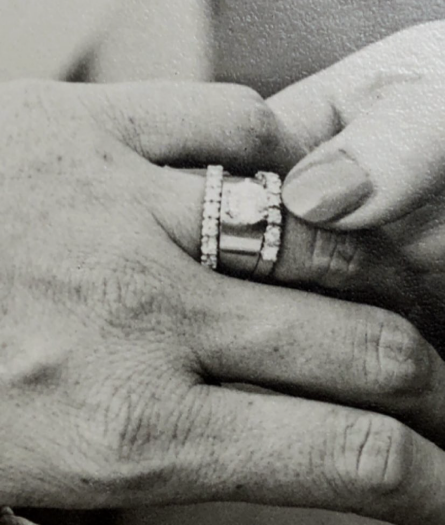 A close-up black-and-white photo of one hand placing a ring with a large stone and diamonds onto another persons ring finger, suggesting a wedding or engagement.